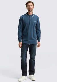 Polo met lange mouwen in donkerblauw met witte kraagdetails. Gecombineerd met donkere spijkerbroek en witte sneakers. Strak ontwerp, casual pasvorm.
