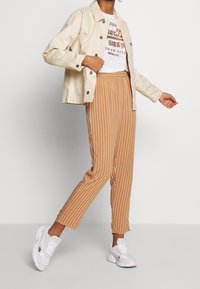 Béžová denimová bunda, bílý grafický tričko a hnědé pruhované kalhoty. Bílé tenisky dokončují outfit, mají texturovaný a přiléhavý design.