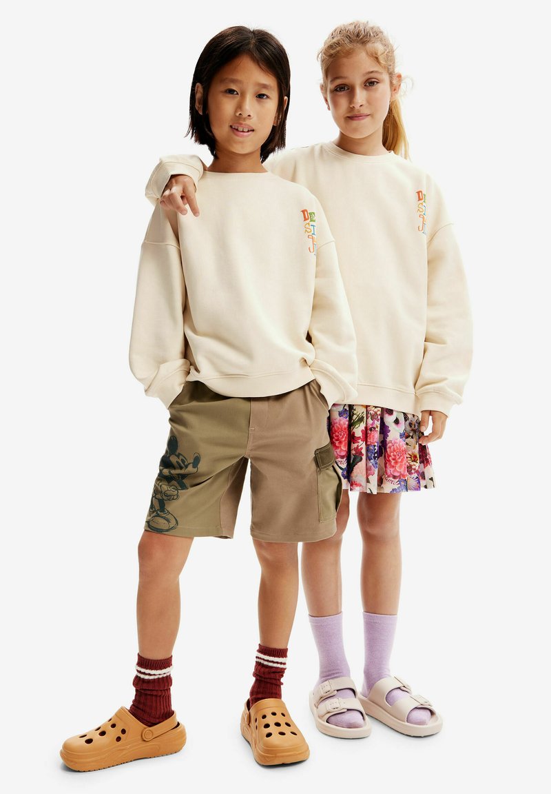 Dos niños están de pie uno al lado del otro, vistiendo sudaderas beige, pantalones cortos caqui con un diseño de Mickey Mouse y faldas estampadas. Llevan calzado casual.