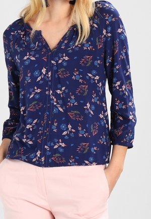 Femme portant un chemisier bleu marine à imprimé floral avec des manches trois-quarts, associé à un pantalon rose clair.