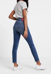 Kobieta w niebieskich jeansach z wysokim stanem, szarej koszulce i białych trampkach, stojąca bokiem na tle białego tła.