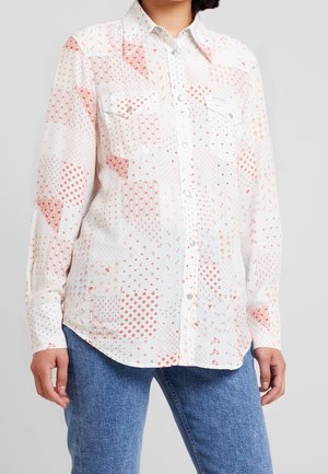 Donna che indossa una camicia bianca con bottoni decorata con motivi geometrici e floreali in rosso, abbinata a jeans in denim blu.