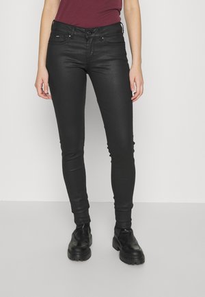 Femme portant un pantalon noir ajusté et des bottes noires épaisses, debout devant un fond blanc uni.