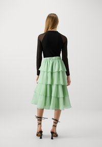 Top negru cu mânecă lungă, asortat cu o fustă din chiffon verde mentă în straturi. Prezintă straturi fluturate și sandale negre cu curele. Textură netedă pe ansamblu.