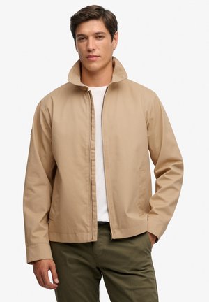 Beige letvægtsjakke med høj krave, lynlås foran og to sidelommer, kombineret med en hvid skjorte og olivengrønne bukser.