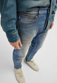 Blue denim jeans with a faded design, featuring a front pocket, contrasting stitching, and a small blue logo tab. Paired with beige sneakers.