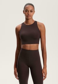 Hnědý sportovní crop top s vysokým výstřihem a bezrukávovým designem, kombinovaný s odpovídajícími legínami s vysokým pasem, vyrobený z hladké, elastické tkaniny.