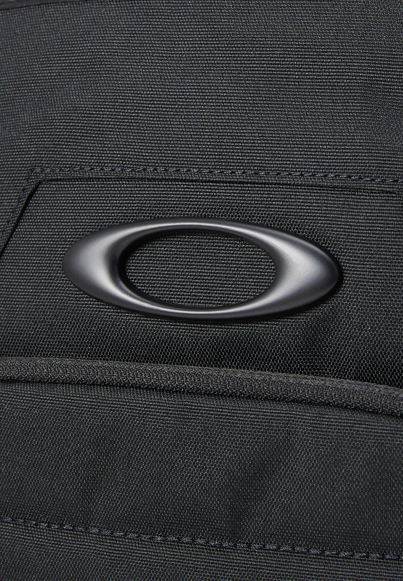 Zwarte stoffen tas met een gestructureerde oppervlakte, voorzien van een opvallend ovaalvormig zwart logo en een bovenvak met stikdetails.