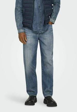 Homme portant un jean en denim bleu clair, des bottes noires, une doudoune sombre et une chemise en denim avec une main dans la poche.