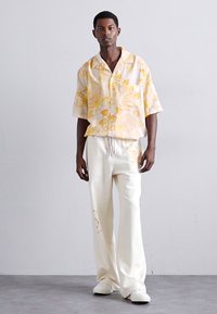 Cămașă cu mânecă scurtă, de culoare galben deschis, cu un model floral, combinată cu pantaloni largi crem și pantofi albi. Material neted și croială lejeră.