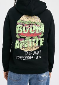 Sudadera negra con una gran gráfica colorida de una hamburguesa de dibujos animados con el texto "¡BOOM APETITO!" y detalles para llevar en la parte inferior.