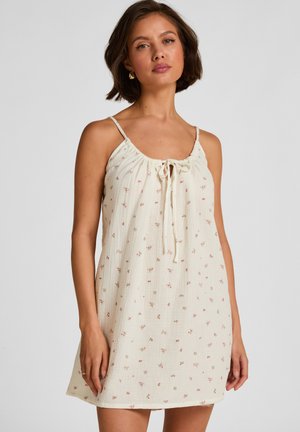 Vrouw draagt een mouwloze, crèmekleurige jurk met dunne bandjes en een klein roze bloemmotief, staand tegen een effen lichte achtergrond.