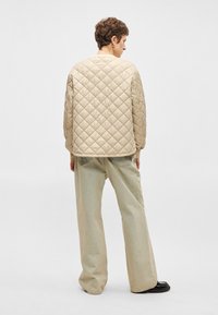 Άτομο που κοιτάζει μακριά φορόντας ένα μπεζ πουκάμισο με επένδυση, ανοιχτόχρωμα παντελόνια με φαρδιά γαζιά και μαύρα παπούτσια, μπροστά σε λευκό φόντο.