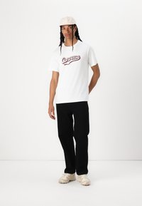 Bijela majica s kratkim rukavima s maroon "Converse" logom, uparena s crnim hlačama i svijetlim cipelama. Jednostavan dizajn i ležeran kroj.
