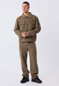 Homme se tenant debout dans un veston et un pantalon à motifs bruns assortis, avec des baskets noires, posé devant un fond blanc uni.