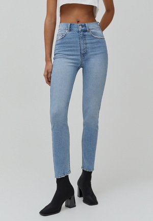 Kvinde iført lyseblå højtaljede slim jeans og sorte ankelstøvler med hæl, stående mod en ensfarvet grå baggrund.