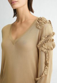 Top beige a maniche lunghe con scollatura a V e dettaglio arricciato testurizzato sulla spalla. Il tessuto sembra leggero e leggermente traslucido.