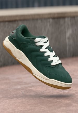 Ciemnozielony sneaker z białymi sznurowadłami i beżową podeszwą wygląda na unoszący się nad różowawą powierzchnią blisko kamiennej ściany.