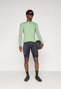 Man wearing green long-sleeve cycling jersey and black shorts, holding black bike helmet, wearing black sunglasses and cycling shoes.