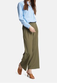Jasnoniebieska koszula z kołnierzykiem i elastycznymi mankietami, w połączeniu z oliwkowo-zielonymi spodniami o szerokich nogawkach i brązowymi sandałami na obcasie z paskami.