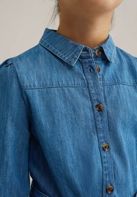 Denim srajca svetlo modre barve z gumbom na sprednji strani, s koničnim ovratnikom in nabranimi rameni. Ima rjave gumbe.