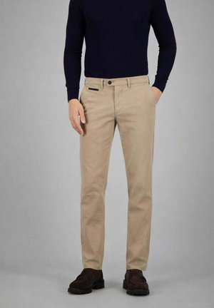 Mann trägt beige Hose mit Seitentaschen, schwarzen Gürtelschlaufen, schwarzem Knopf, dunklen Loafers und einem enganliegenden schwarzen Langarmoberteil vor grauem Hintergrund.