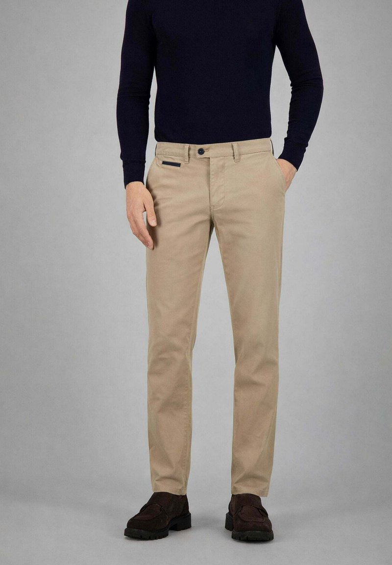 Uomo che indossa pantaloni beige con tasche laterali, passanti per cintura neri, bottone nero, mocassini scuri e top nero aderente a maniche lunghe su sfondo grigio.
