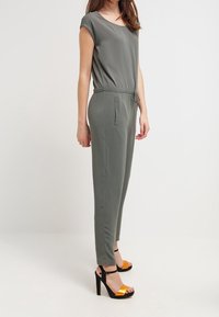 Zielono-oliwkowy kombinezon z krótkimi rękawami, elastyczną talią i bocznymi kieszeniami, w połączeniu z czarnymi i pomarańczowymi sandałami na obcasie.