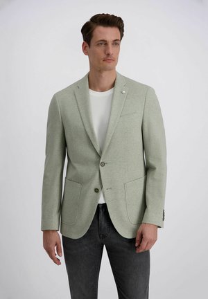 Man draagt lichtgrijze blazer over wit overhemd en donkere spijkerbroek, staand tegen een effen lichte achtergrond.