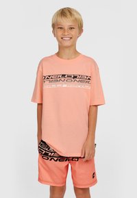 Lichtroze katoenen t-shirt met vetgedrukte zwarte tekst en logo-ontwerp, gecombineerd met koraalkleurige shorts met zwarte branding. Korte mouwen, relaxte pasvorm.