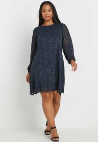 Vestido de encaje azul marino con mangas largas transparentes, dobladillo con forma de concha y escote redondo. Presenta un patrón floral y acentos de lentejuelas.