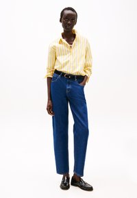 Camicia a righe gialle e bianche, pantaloni dritti in denim blu, cintura nera e mocassini, su uno sfondo bianco semplice.