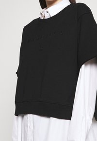 Svart cropped sweatshirt med upphöjd text "MARGIELA 6" och ribbad nederkant. Bärs över en vit skjorta med krage. Mjuk textur.