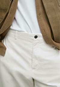 Lichtgekleurde linnen broek met een gladde textuur, voorzien van een zwarte knoopsluiting, gedragen met een bruine jas en een wit shirt.