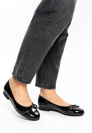 Black patent leather ballet flats with a rounded toe and small bow detail. The sole is slightly elevated, paired with gray denim pants.