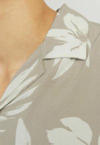 Jasnoszara koszula z kołnierzykiem z dużymi, białymi wzorami kwiatowymi. Gładka tekstura materiału z delikatnym połyskiem. Zbliżenie na kołnierz i dekolt.