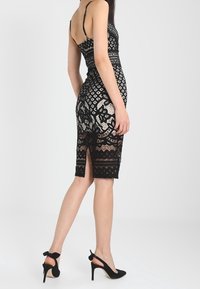 Robe en dentelle noire au design ajusté, avec une fente au dos et des motifs géométriques. Portée avec des talons noirs ornés de nœuds.