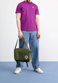 Lila bomulls-T-shirt med en liten vit logotyp, kombinerad med ljusblå, löst sittande jeans. Oljefärgad budväska med en rund lapp.