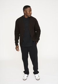 Ruskang cardigan yang terbuat dari rajutan coklat di atas kaos hitam, dipasangkan dengan celana hitam dan sneakers putih dengan aksen abu-abu. Desain sederhana, penutupan tombol.