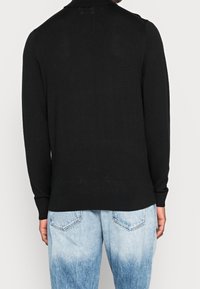 Pull noir en maille avec col, manches longues et ourlet ajusté, associé à un jean bleu clair avec un dégradé progressif.