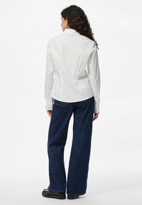 Camicia bianca aderente con colletto, cuciture posteriori e maniche lunghe, abbinata a jeans azzurri a gamba larga e scarpe nere.