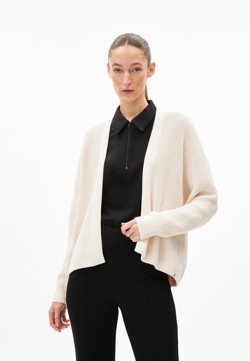 Cardigan beige clair en maille texturée, avec un devant ouvert, des manches côtelées et une coupe décontractée, associé à un top noir.