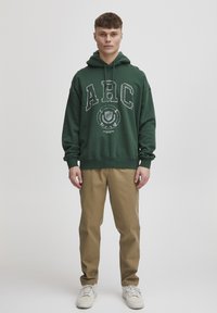 Hanorac verde cu glugă, cu logo "ARC", croială lejeră, din material de bumbac. Asortat cu pantaloni chino bej deschis și adidași albi.