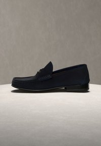 Ναυτικό μπλε σουέντ ανδρικό μοκασίνι με χαμηλό τακούνι, ραφή μοκασίνου και διακοσμητικό μεταλλικό στοιχείο στην κορυφή, τοποθετημένο σε ανοιχτή επιφάνεια.