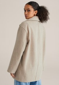 Ženska kodrastih las v nizek čop, oblečena v bež volneni plašč in modre kavbojke, gleda preko ramena na enobarvnem ozadju.