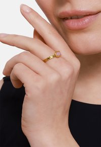 Bague en or torsadée avec une pierre carrée de couleur rose clair. La main est montrée près du visage, avec une peau lisse et un fond neutre.