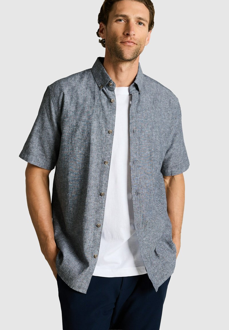 Homme portant une chemise bleue à manches courtes à motifs, boutonnée, par-dessus un t-shirt blanc et un pantalon foncé, debout les mains dans les poches.