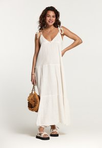 Robe maxi blanche sans manches avec un décolleté en V, texture froncée et bretelles à nouer. Assortie à un sac marron texturé et des sandales légères.
