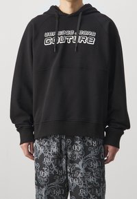 Černá mikina s kapucí s výrazným bílým logem "VERSACE JEANS COUTURE", vyrobená z měkké látky s volným střihem a žebrovanými manžetami.