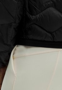 Acercamiento del dobladillo de una chaqueta negra acolchada sobre unas mallas beige de cintura alta con detalles de costura visibles y una mano descansando a un lado.